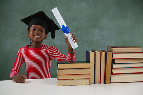 Portrait of schoolgirl in mortar board holding certificate against chalkboard