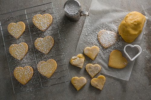 Overhead of raw heart shape cookies on baking tray with flour shaker strainer, cookie cutter and wax paper