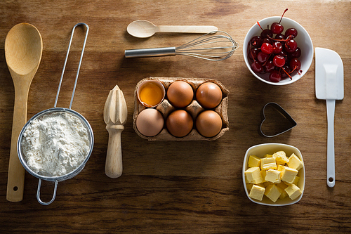 Over head view of eggs, butter cubes, cherries, sieve and whisk on a wooden table