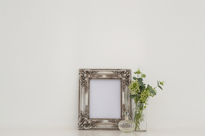 Picture frame, perfume bottle and flora on table against white wall