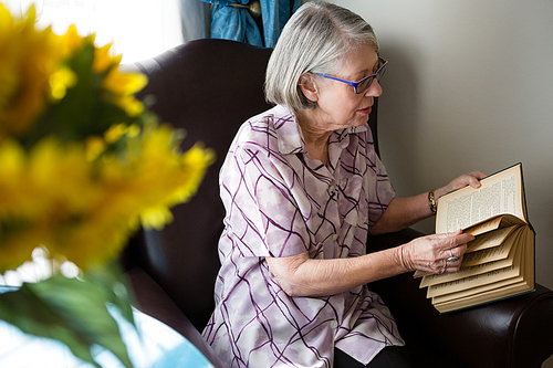 Senior woman reading book while sitting on arm chair in nursing home