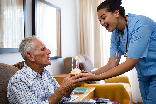 Senior man blowing candles of cup cake being held by female doctor in nursing home