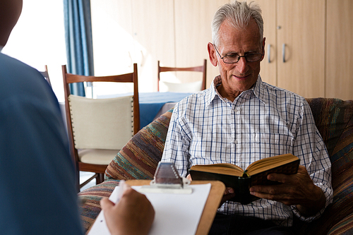 Female doctor writing on paper while senior man reading book in nursing home