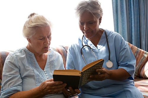 Doctor with senior woman reading book while sitting on sofa in nursing home