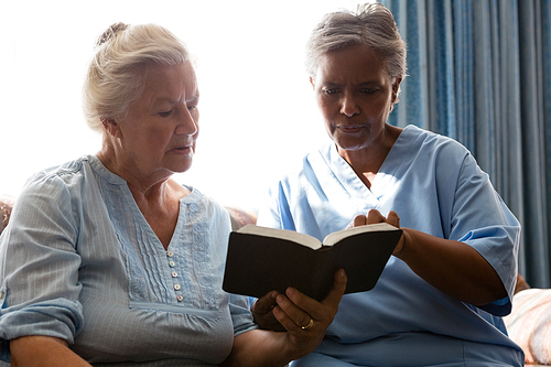 Doctor with senior woman reading book while sitting on sofa in retirement home