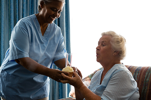 Senior woman blowing candles of cup cake being held by nurse in nursing home