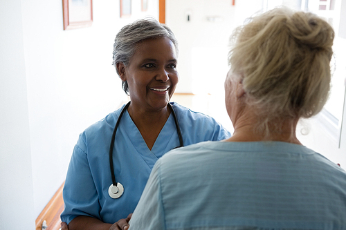 Senior woman and nurse talking while standing in corridor at nursing home
