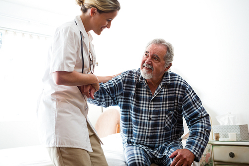 Female doctor assisting senior man in sitting on bed at nursing home