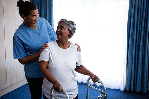 Smiling nurse assisting senior woman in walking with walker at nursing home