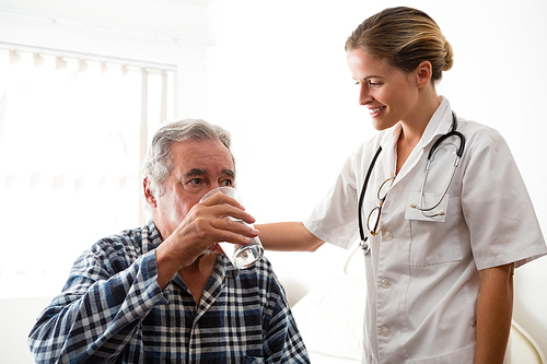 Female doctor standing by senior man drinking water while sitting at nursing home