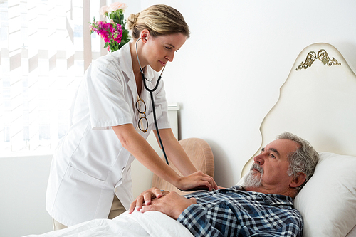 Female doctor listening to heartbeats of senior man lying on bed in nursing home