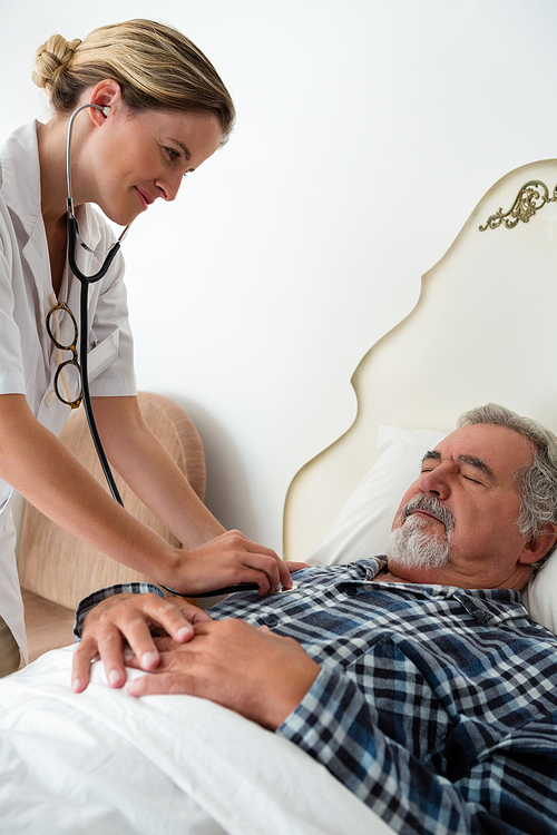 Female doctor listening to heartbeats of senior man sleeping on bed in retirement home