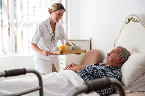 Female doctor serving food to senior patient relaxing on bed in nursing home