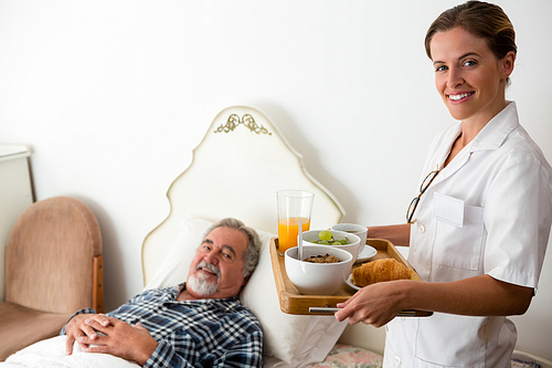 Portrait of female doctor serving food to senior patient relaxing on bed in retirement home