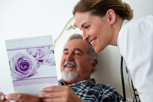 Female doctor showing get well card to patient relaxing on bed in retirement home