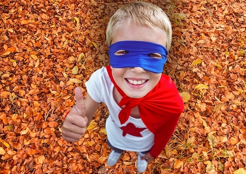 Composite image of smiling kid in red cape and blue mask showing thumbs up during autumn