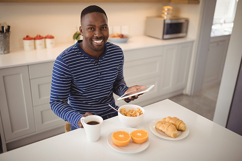 Portrait of smiling man using a digital tablet while having breakfast in kitchen at home