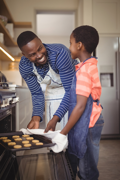 Smiling father taking tray of fresh cookies out of oven with son in kitchen at home