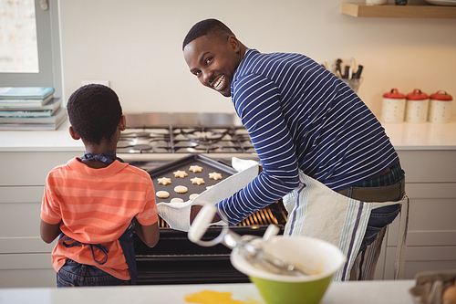 Smiling father taking tray of fresh cookies out of oven with son in kitchen at home