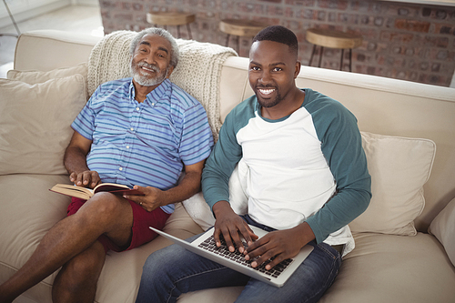 Portrait of smiling father and son sitting on sofa with laptop and book in living room