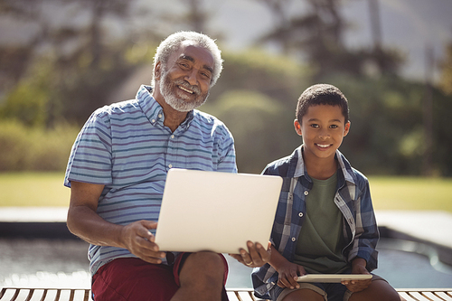 Portrait of smiling grandfather and grandson sitting together on bench with laptop and digital tablet