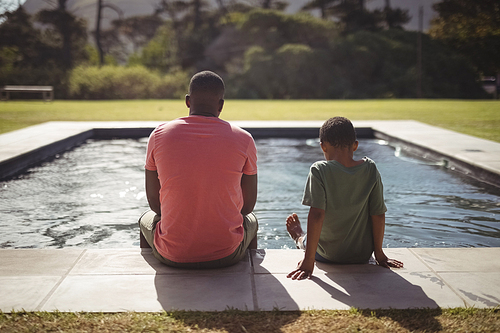Rear view of father and son sitting on edge of swimming pool