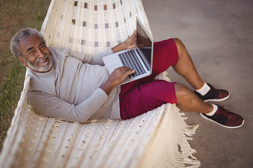 Portrait of smiling senior man using laptop while relaxing on hammock