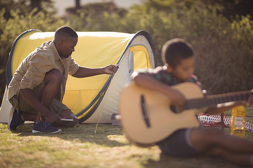 Boy playing guitar while father setting up a tent in park on a sunny day