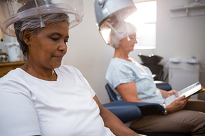 Senior women reading magazie while sitting under hair steamer in salon