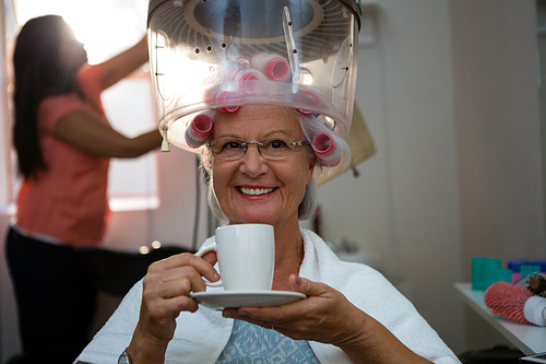 Portrait of senior woman having drink while sitting under hair steamer in salon