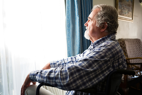 thoughtful senior man looking through window while sitting on . in retirement home
