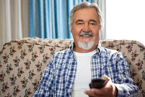 Portrait of smiling senior man operating remote while sitting on sofa in nursing home