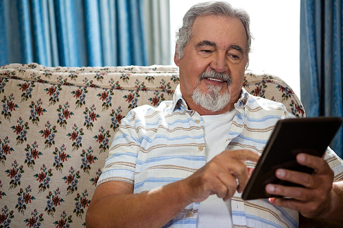 Smiling senior man using digital tablet while sitting on sofa in nursing home