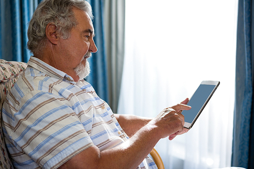 Side view of senior man using digital tablet while sitting on sofa in nursing home