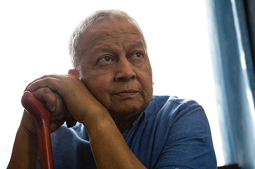 Thoughtful senior man holding walking cane while sitting by window in nursing home
