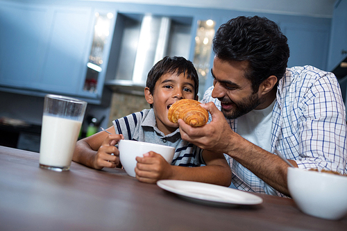 Man feeding croissant to son while having breakfast at home