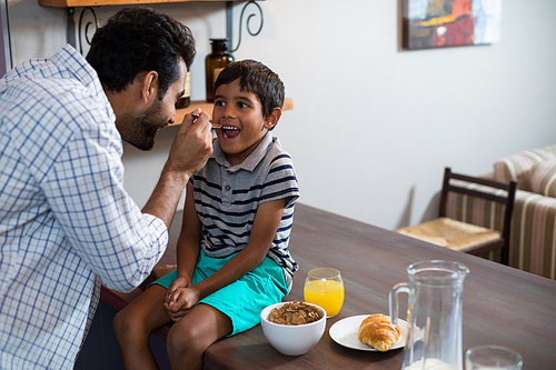 Happy father feeding cereal breakfast to son sitting on table at home