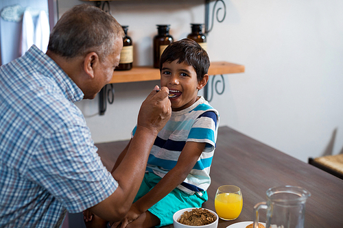 Happy grandfather feeding grandson sitting on table at home