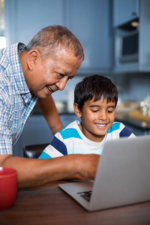 Smiling grandfather assisting grandson using laptop in kitchen at home