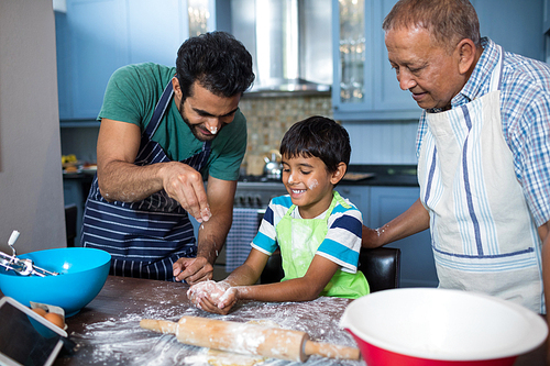 Father sprinkling flour on sons hand while preparing food with grandfather in kitchen at home