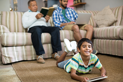 Smiling boy using tablet lying on carpet with father and grandfather sitting on sofa in background