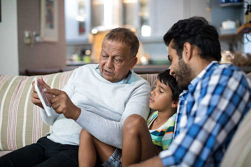 Grandfather showing tablet to grandson sitting with father on sofa in living room at home