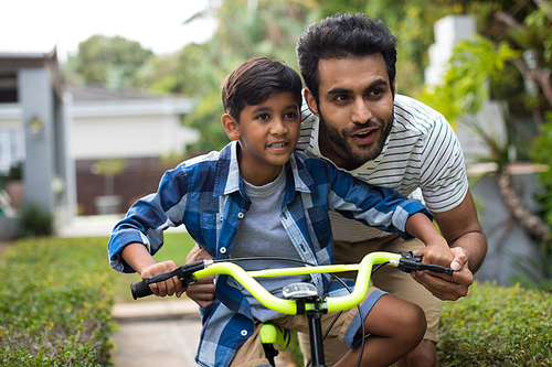 Father looking away while assisting son for cycling in yard