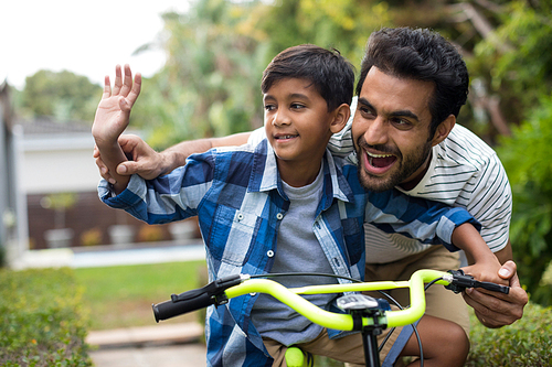 Father and son waving hand while cycling in yard