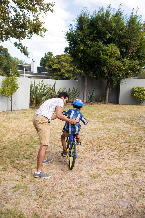 Rear view of father assisting son while riding bicycle on field in yard