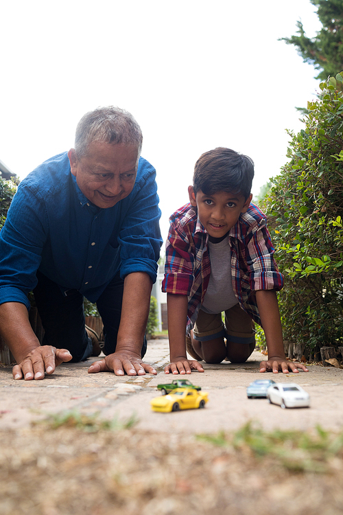 Boy and grandfather playing with toy cars while kneeling on pavement in yard against sky
