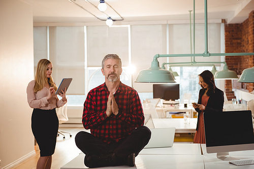 Male executive meditating on table while colleague working in background at office