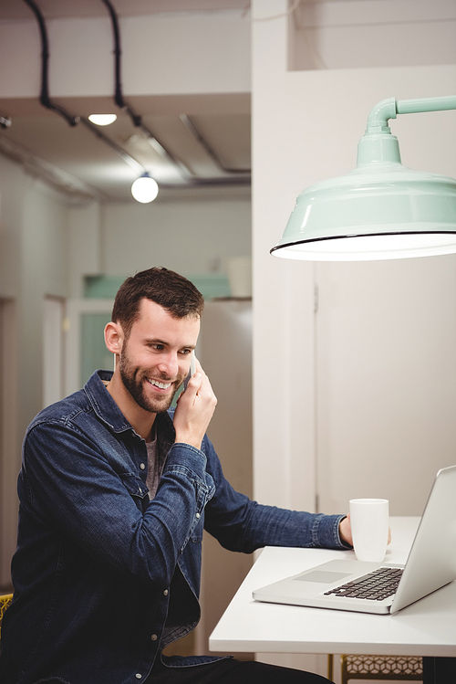 Smiling male executive talking on mobile phone while having coffee in office