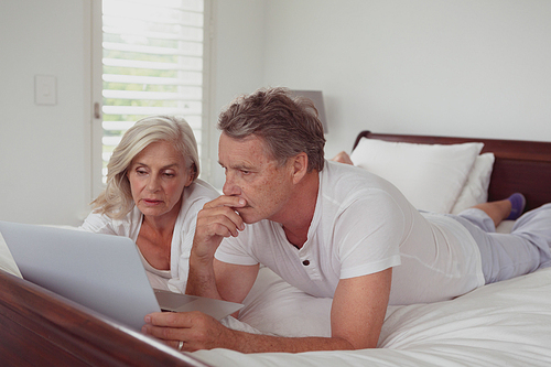 Front view of active senior Caucasian couple using laptop while lying on bed in bedroom at comfortable home