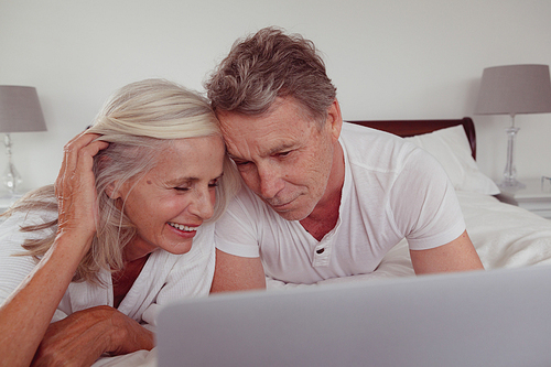 Front view of active senior Caucasian couple using laptop while lying on bed in bedroom at comfortable home